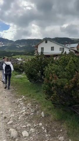 The video shows a group of three people hiking on a dirt path through a mountainous landscape. The path is lined with green vegetation and scattered rocks. In the background, there is a large, white building with a red roof and several windows. Beyond the building, snow-capped mountains rise under a cloudy sky. One person in the group is wearing a backpack, and another is wearing a white hoodie. The group is walking away from the camera towards the mountains and the building. The scene appears to be a hiking trail leading to a mountain lodge.