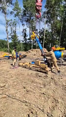 In an outdoor construction setting, a crane with a red hook is lifting a piece of heavy industrial equipment that is being guided by two workers. The equipment is blue and black and has yellow accents. One worker, wearing a tan jumpsuit and an orange hard hat, is standing to the right of the equipment, holding it steady. Another worker, in a blue jumpsuit and a black hard hat with a yellow vest, is standing to the left of the equipment, also guiding it. The equipment is suspended over a wooden pallet. In the background, there are large blue pipes, mounds of dirt, and trees under a blue sky. The ground is mostly dirt and gravel.