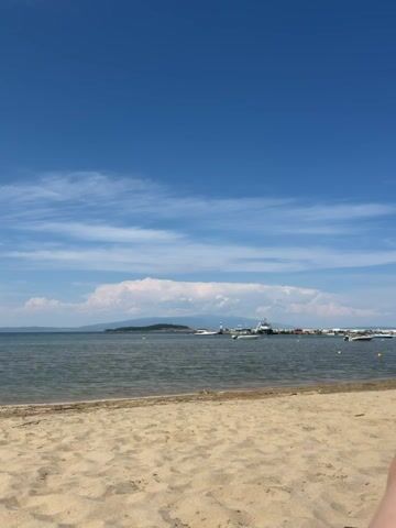 The video shows a view of a sandy beach and the sea on a clear day. The sky is blue with some wispy white clouds. The sea is calm and reflects the sky. In the distance, there is a landmass with hills and trees. There is a small port with boats and a lighthouse. The beach is made of sand and appears to be empty, except for a small part of a person's leg visible in the bottom right corner. The video is shot from a stationary position.