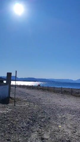The video shows a scenic view of a beach on a sunny day. The sun is shining brightly in a clear blue sky. There is a dirt path in the foreground leading towards the ocean. A low fence runs along the edge of the path, separating it from the beach. In the distance, the ocean stretches out, with gentle waves lapping at the shore. Hills or mountains can be seen on the horizon across the water. Several people are visible on the beach, some walking and others possibly sitting or standing. There is also a white structure on the left side of the frame, possibly a building or a wall, with a post and a sign next to it. A palm tree is visible to the left of the structure. The overall impression is one of a peaceful and beautiful coastal landscape.