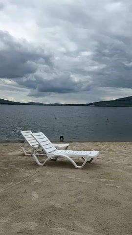 A person is bending over at the edge of a body of water. Two white lounge chairs are in the foreground, on a sandy or pebbly surface. The water is dark and appears to be a lake or sea. The sky is cloudy and gray, with hills visible in the distance.