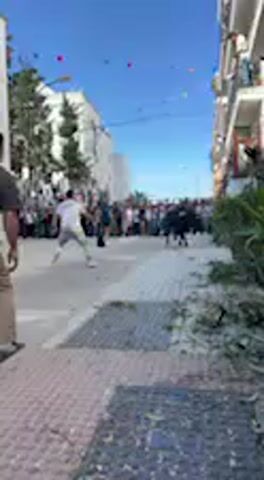 The video captures a bull-running event taking place on a sunny day in what appears to be a town street in Vejer de la Frontera, Spain. A large, dark-colored bull is the central subject, initially running down a relatively narrow street lined with multi-story buildings on both sides.

In the foreground, a person quickly steps away as the bull moves past. Numerous people are visible further down the street in the background, and spectators can be seen on the balconies of the buildings, watching the event unfold. The street itself has a paved sidewalk and a road surface, with some green foliage, possibly small bushes, along the building walls.

Overhead, colorful decorative flags or bunting are strung across the street, suggesting a festive occasion. The sky is bright blue and clear, indicating good weather. As the video progresses, the bull pauses near a bush on the sidewalk, appearing to be momentarily stationary before potentially continuing its run. The architecture of the buildings is typical of a Spanish town. There is no visible text in the video.