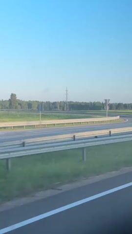 This video captures a view from a vehicle traveling along a highway in Medemciems, Latvia. The scene is dominated by the clear blue sky and a lush green landscape. The highway itself is prominent, with its asphalt lanes marked by white lines and bordered by metal guardrails.

Several objects and signs are visible along the roadside. In the distance, a tall electricity pylon stands against the horizon, suggesting the presence of power infrastructure. There are also large, empty overhead signs that are likely used for advertising or directional information, though no text is discernible on them. Closer to the road, traffic signs are present. One sign displays three upward-pointing arrows and the number "200", possibly indicating lane usage or distance to a point. Another sign is a circular red sign with a horizontal white bar, which is a universal "no entry" or "do not enter" sign.

The time of day appears to be late afternoon or early evening, given the warm, soft light bathing the landscape and the long shadows cast by the guardrails. The weather seems to be fair and clear, with no clouds in the sky. The overall impression is one of peaceful travel through a rural or semi-rural area, with the infrastructure of a modern highway integrated into the natural surroundings. There are no people visible in the frame, and the focus is on the road and the environment. The overall mood is tranquil and observational.