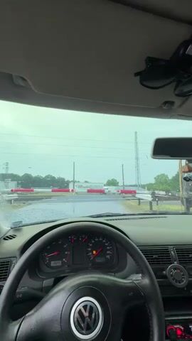 The video captures the perspective from inside a car, showing the dashboard and windshield. The windshield wipers are active, indicating rainy weather. Outside, a railroad crossing barrier is down, with red and white striped poles blocking traffic. A train, primarily blue with red accents and the word "BULMARKET" visible on its side, passes across the tracks from left to right. Another train, gray and red, follows behind the first. The sky is overcast.