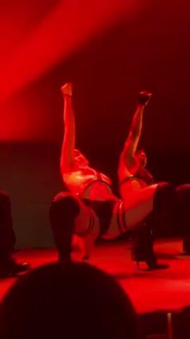 Here is a concise description of the video:

The video shows a group of dancers performing on stage under red lighting. The central figure is a dancer wearing a revealing outfit consisting of a black bikini-style bottom, a clear halter top, and high-heeled black boots. The dancer is flanked by other performers, some of whom appear to be male, and all are moving in unison. The performance is energetic, with the central dancer raising an arm in the air at times. The background is dark.