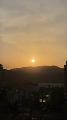 The media file captures a serene sunset over a mountainous landscape, likely in Les Amplaries, Spain. The sun, a bright orb, is positioned just above the crest of a silhouetted mountain range, casting a warm, golden glow across the sky. Wisps of clouds are visible, tinged with the same orange and yellow hues of the setting sun, suggesting a clear or partly cloudy evening.

In the foreground, the silhouettes of buildings and trees are visible, indicating an urban or semi-urban environment at the base of the mountains. The architecture of the buildings suggests a resort or residential area, with one tall, multi-story building on the left and a shorter structure on the right. Palm trees are also discernible, adding to the Mediterranean feel of the location. The scene is quiet and peaceful, evoking a sense of tranquility as day transitions into night. The time of day is clearly sunset, characterized by the low position of the sun and the warm, diffused light. There is no visible text within the media file.