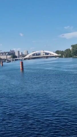 The video features a view from a moving boat, looking out at a wide blue river with gentle ripples. In the distance, a modern white arched bridge spans the river. To the left, a city skyline with various buildings, including what appears to be a distinctive contemporary building with a metallic, angular roof, is visible. Several cylindrical buoys, red and green striped, are anchored in the river. The sky is clear and light blue. The boat's railing or window edge is briefly visible in the bottom left as the camera pans slightly.