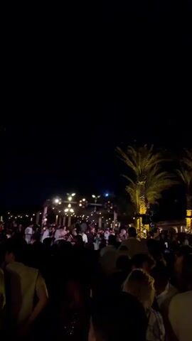 This is a nighttime outdoor scene at a crowded event. A large group of people are gathered, with many standing and some appearing to be dancing. The background features palm trees and structures adorned with numerous string lights and overhead lights, suggesting a festive or event-like atmosphere. Some individuals are holding up phones, possibly to record or take pictures. The lighting is dim, creating a nightlife ambiance.