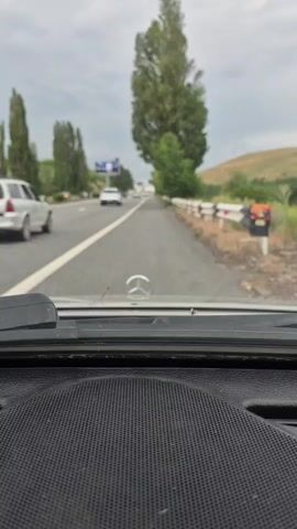 The video shows a view from inside a car driving on a highway. The camera is focused on the dashboard and the windshield. The road ahead is visible, with other cars driving in the same direction. Tall trees and green hills are on the right side of the road. The sky is overcast.