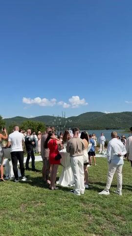 The video shows an outdoor wedding ceremony taking place on a sunny day with a clear blue sky. A body of water with forested hills in the background serves as the backdrop. Guests are gathered on a grassy lawn, some standing around tables with drinks, while others are positioned to watch the ceremony. The ceremony area is marked by a white aisle runner and chairs. Several people are gathered at the front, presumably the wedding party and officiant. The camera focuses on different groups of people and shifts perspectives throughout the brief recording, capturing the atmosphere of the event.