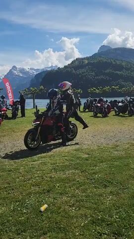 The video shows a group of people on motorcycles parked on a grassy field, with a scenic backdrop of a lake, mountains, and a partly cloudy sky.  Two people, wearing helmets and protective riding gear, are seated on a red motorcycle that starts to drive away. Other motorcycles are parked nearby, some with people standing around them. A red banner with white text is visible. The terrain is mostly green grass with some bare patches. A discarded beverage can is on the ground.
