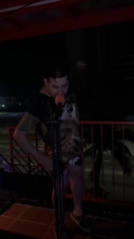 A man is standing on a small stage or platform on a dark night. He is wearing a colorful shirt, shorts, and gray Crocs. He has tattoos on his arms, and a cigarette in his mouth. There is a microphone stand in front of him, with a red microphone on top. The stage is made of red metal bars and red tiles. In the background, there is a road and a few buildings. The lights on the stage change color, cycling through red and green. The man gestures to the side, then dances, bending forward and waving his arms.