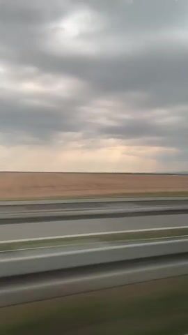 This is a video recording of a landscape viewed from a moving vehicle, likely a train or car, as indicated by the blurred motion of the foreground. The scene is dominated by a vast, harvested agricultural field that stretches to the horizon. The field is a uniform, light brown color, suggesting it is either stubble or recently tilled earth. Subtle lines and patterns are visible across the field, indicating the direction of agricultural work.

A narrow strip of green vegetation runs along the edge of the field closest to the viewer. In the foreground, two parallel gray lines with a white stripe in between suggest a road or railway. The sky is overcast with thick, dark gray clouds, but a lighter, almost hazy band of illumination is visible near the horizon, hinting at the presence of the sun behind the clouds, possibly indicating late afternoon or early evening. The overall mood is subdued and perhaps a little melancholic due to the cloudy sky and vast, empty field.

There is no visible text in the video. The setting appears to be rural, likely in Serbia given the provided location context of Indjija Municipality. The activity is passive observation of the passing landscape during travel.
