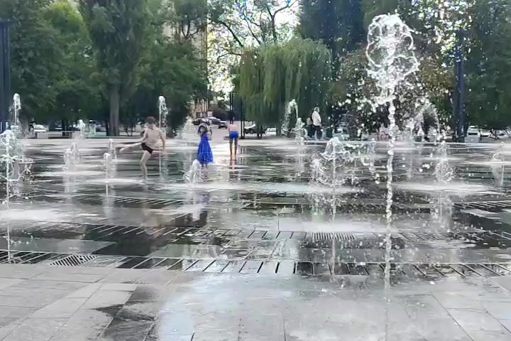 The video shows a public square with a water fountain feature. Several people are present, including at least two children and several adults in the background. Two children are in the foreground, one in a blue dress and one in swim trunks. They are walking through the water jets of the fountain. The ground is paved with square tiles, and there are trees and buildings in the background. The water feature consists of multiple jets of water spraying from the ground in a synchronized pattern. The scene appears to be a warm day, as people are dressed in light clothing and some are barefoot.
