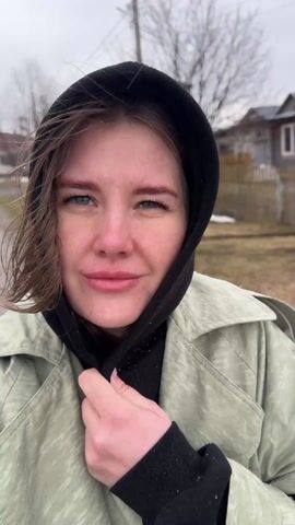 A young woman with light brown hair and blue eyes stands outside while it snows. She wears a black hoodie under a light green trench coat. She looks up at the falling snow, closes her eyes, and then smiles at the camera. A small, wooden house and a wooden fence are visible in the background. The ground is covered in patches of grass and snow.