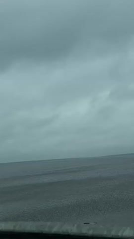 The video shows a wide expanse of water under a gray, overcast sky. The water has subtle ripples, and the horizon line is visible in the distance with a dark strip of land. A metallic barrier with rivets runs along the bottom of the frame. There are no discernible people, objects, or actions beyond the view of the landscape.