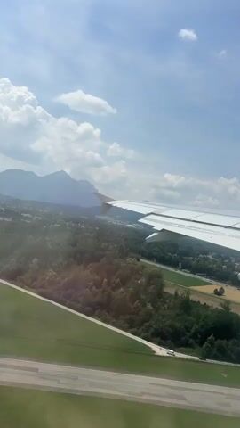 This video captures an aerial view from an airplane window, likely during takeoff or landing, over the outskirts of Siezenheim, Austria. The scene is dominated by the white wing of the aircraft, extending prominently into the frame from the right. Below, a landscape unfolds, characterized by a patchwork of green forests, agricultural fields in various shades of brown and gold, and a winding highway with visible traffic. In the distance, a mountain range is silhouetted against a bright blue sky dotted with fluffy white clouds.

The time of day appears to be daytime, with ample sunlight illuminating the scene, suggesting clear or partly cloudy weather. The presence of the highway, fields, and a few scattered buildings indicate a semi-rural or suburban environment. The overall impression is one of serene transition, as the aircraft moves through the air, offering a panoramic vista of the Austrian countryside. No visible text is present in the video.
