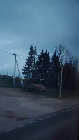 A nighttime or dusk video shows a rural roadside scene. The sky is dark blue, indicating a low light condition. On the left side of the frame, there are tall evergreen trees and some deciduous trees with bare branches. A power pole with lines is visible. In the foreground, there is a paved road with a white line marking the lane. A truck with a brown cargo bed is parked to the left of the road, partially obscured by trees. Further down the road, on the right side, a traffic sign with a red circle and a diagonal line through it is visible. The scene appears to be in a natural, undeveloped area.