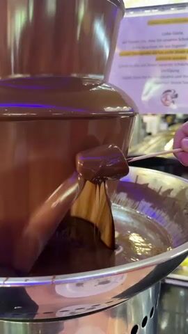 The video shows a chocolate fountain in operation, with melted chocolate flowing down its tiers. A person's hand, visible on the right side of the frame, is holding a skewer with a marshmallow on it, dipping it into the chocolate. The marshmallow is being coated in the flowing chocolate. In the background, there is a sign with German text and what appears to be decorative lighting. The fountain is made of dark brown material, and the basin at the bottom is filled with chocolate.