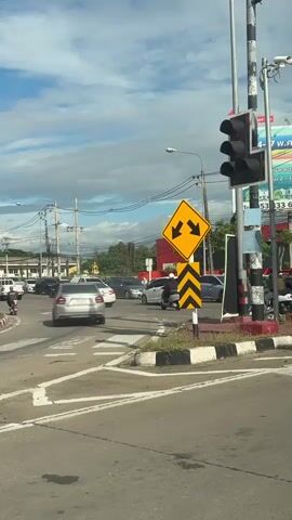 The media shows a traffic intersection in Tha Sala, Thailand. In the foreground, a traffic light and a diamond-shaped yellow sign with two black arrows pointing in opposite directions are visible, indicating a lane split. The sign is mounted on a black and yellow striped pole. In the background, several cars and a scooter are navigating the intersection under a partly cloudy sky. The sun is likely out as indicated by the brightness and shadows. The overall scene depicts a typical urban traffic environment. There is some visible text on a sign in the background, but it is too blurry to read clearly.