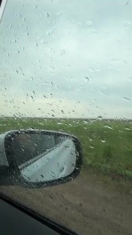 The video shows a view from inside a car on a rainy day. Raindrops cover the side window, partially obscuring the view of a green field and a dirt road. The car's side mirror is visible, reflecting the surrounding scenery. The sky is overcast. The car appears to be moving slowly, as the landscape outside remains relatively static.