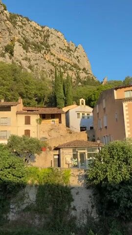 A still, vertical video captures a view of houses nestled against a steep, rocky mountainside under a clear blue sky. The houses, built in a traditional Mediterranean style with terracotta roofs and stucco walls, appear to be on a slope, with vegetation, including trees and ivy, growing around them. The sun appears to be low in the sky, casting warm light on the scene, suggesting either early morning or late afternoon. The camera is static, and there are no people or discernible actions taking place. The environment is natural, with the prominent mountain and abundant greenery creating a picturesque, tranquil setting.