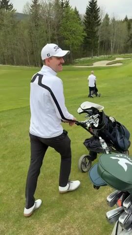 A man is walking on a golf course pulling a three-wheeled golf bag trolley. He is wearing a white jacket with black accents, black trousers, a white golf cap, and white golf shoes. A golf bag is visible in the foreground. In the background is a green golf course with trees and sand traps. Another person is visible in the distance. The sky is overcast.