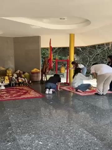 A group of people are on their knees in a temple-like setting, with one person striking a large gold bell hanging on a red and gold stand. There is a red patterned rug under them. Behind the people is a tableau of small figurines and statues. In the background, one can see greenery and other architectural elements of the temple.
