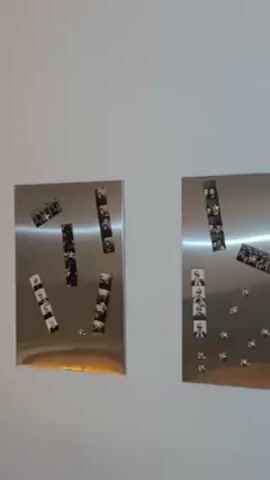 The video shows two square metal boards hanging on a white wall. Each board has several strips of black and white film negatives attached to it with small round magnets. The film negatives appear to depict portraits of people. The lighting in the room is bright.