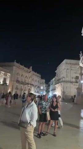 The video is a shaky, blurred night-time scene of a public square with people walking around and a street performer playing a guitar. The buildings surrounding the square are illuminated, and the sky is dark. In the foreground, a woman in a white jacket is walking away from the camera. To her right, a group of people are standing and talking. Further into the square, a man is playing a guitar while sitting on the ground. A bicycle is leaning against a lamppost next to him, and there are some items on the ground in front of him, possibly for donations. Another person in a red shirt is standing behind him. The camera pans and zooms slightly throughout the short clip, making it difficult to discern clear details about the individuals.