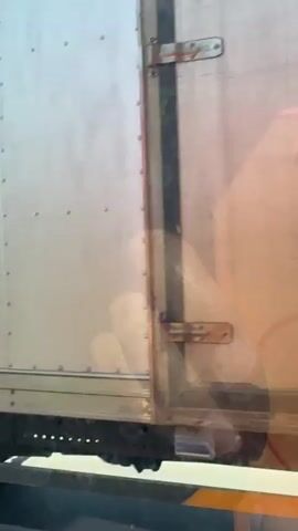 The video shows a close-up view of the side of a white truck, viewed from a moving vehicle. The truck appears to be in motion, as indicated by the slight blur of the background and the movement of the truck itself in relation to the camera.

The subjects visible are the side of the truck, which is made of a metallic material and has several rivets along its surface. On the truck's door, there are two prominent decals: one is a stylized dragon's head, depicted with vibrant colors and intricate details, and the other is a circular emblem with text. The dragon decal is on the rear section of the truck's side, while the circular emblem is further forward.

There are also reflections on the truck's surface, suggesting it is daytime and the sun is out, though the overall lighting is somewhat diffused. A partial view of a person's hand pressing against the inside of the truck's door is visible through a semi-transparent panel. The hand appears to be pressing from the inside, with the fingers slightly distorted by the material. A yellow object with a yellow tie is also attached to the truck's exterior.

In the lower part of the frame, a yellow and white striped safety line is visible, indicating the truck is on a road. The background is blurred, but it suggests an outdoor environment, possibly a highway or a road in a developed area, given the presence of the truck and the road markings.

The activity is primarily observational, focusing on the details of the truck's exterior and the reflections on its surface. There are no strong emotional cues conveyed, as the focus is on inanimate objects and a brief glimpse of a hand.

The visible text on the circular emblem is difficult to read clearly due to its small size and the angle, but it appears to be a type of seal or certification. The general location context provided is Hòa Đa, Vietnam. The time of day is likely daytime due to the lighting. The weather appears to be clear enough for outdoor travel and reflections to be visible.