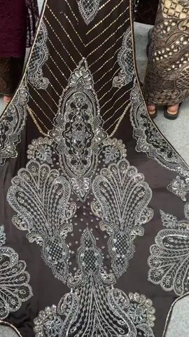 The video shows a close-up, top-down view of the back of a traditional Indonesian wedding dress on a mannequin. The dress is primarily black with elaborate silver and gold beadwork and sequin embellishments. The skirt is long and flowing, featuring large, ornate paisley-like patterns and diagonal stripes on the train. The upper part of the dress is also heavily decorated with sequins and beadwork. The mannequin is adorned with a traditional Javanese bridal headdress, which includes golden hair ornaments and floral elements. Around the mannequin, several people are visible, mostly from the chest down, wearing formal attire, including a maroon top, a dark patterned skirt, and a light-colored hijab. The environment appears to be an indoor event, possibly a wedding reception, with some decor visible in the background. The camera pans slightly, offering a glimpse of other guests and the general setting.