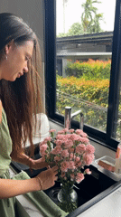 Woman arranges pink flowers in kitchen, Thailand morning scene