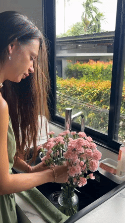 Woman arranges pink flowers in kitchen, Thailand morning scene
