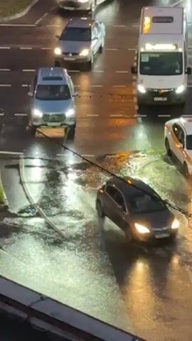 The video shows a view from above of cars driving on a wet road at night. The road appears to be an intersection, with multiple lanes and traffic lights. Cars are seen driving, turning, and stopping. One car, a silver sedan, appears to be making a turn and splashing water. The lighting suggests streetlights and the headlights of the vehicles. The overall scene is one of normal urban traffic during nighttime conditions with rain.