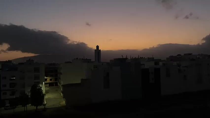 The video captures a cityscape at dusk. The sky is a gradient of deep purple, orange, and yellow, with dark clouds partially obscuring a mountain range in the distance. A tall, slender minaret of a mosque stands prominently against the twilight sky, surrounded by the rooftops of various buildings. The buildings are mostly low-rise, appearing to be apartments or residential blocks, with some windows illuminated by interior lights. A street with a few parked cars and trees is visible in the foreground, also dimly lit by streetlights. The overall scene is serene and tranquil, evoking a sense of the transition from day to night in an urban environment.