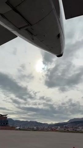 The camera is situated underneath the wing of an airplane, looking out at a city built on a hillside. The city consists of many closely spaced buildings, mostly red and brown. Mountains are visible in the background. The sky is cloudy, with the sun just barely visible through the clouds. There is an orange traffic cone in the foreground. Airport vehicles are moving on the tarmac.