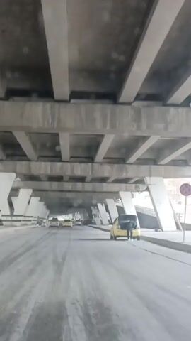 The video shows a car driving on a road under an overpass. The overpass is made of concrete and has a grid-like structure. Several other cars are on the road, some of which are yellow taxis. A man is standing near a yellow taxi on the right side of the road with the trunk open. The road surface is grey and appears slightly dusty. The overall lighting is dim due to the shade from the overpass.