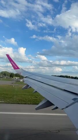 The video shows the wing of an airplane as it taxis on a runway. The wing is gray with a pink tip and the Wizz Air logo. The sky is blue with white clouds. Green grass and trees are visible on either side of the runway. The plane appears to be moving forward slowly.