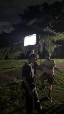 The video shows a young man at night in a grassy area with a dark-colored car in front of him. The man is wearing a dark blue t-shirt and khaki shorts. He has curly dark hair. He is holding a device in his hands, possibly a remote control. He is looking at the camera and talking. The car is a classic model, and the headlights are on. The background includes trees and some distant lights. It is a dark, cloudy night.