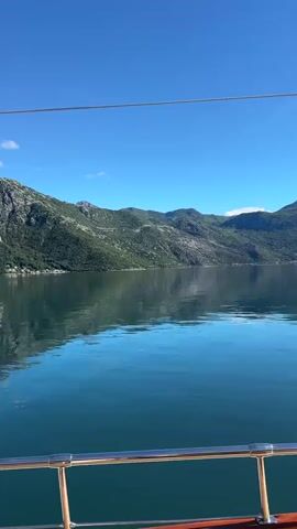 The video presents a calm, steady shot of a tranquil bay surrounded by mountains, likely taken from a boat. The water is mostly still, reflecting the green, forested mountains and the clear blue sky. The sunlight is strong, creating a bright, sunny day effect. The camera is positioned at the edge of the boat, as a stainless-steel handrail is visible along the lower edge of the frame. A small town or settlement is visible along the shoreline in the distance. The atmosphere is peaceful and scenic.