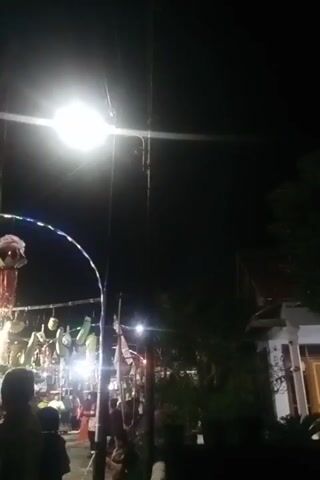 The video opens with a view of a street at night, illuminated by a bright overhead light. In the lower left quadrant, a person's back is visible, facing away from the camera. They appear to be standing in a crowd of people. Partially visible in the background are decorations, including what looks like an arched structure adorned with lights and hanging items. Further back, there are more people and stalls, suggesting a market or festival. In the upper portion of the frame, fireworks briefly appear and dissipate. The scene is dark with artificial lighting from the streetlamp and decorations. The overall impression is of a lively nighttime gathering.