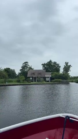 A wide shot shows a dark wooden house with a thatched roof situated on a small island in the middle of a calm body of water. Trees surround the house. The sky is overcast. The foreground shows the edge of a red boat. The scene appears to be a still shot, as there is no discernible movement.