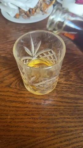 The video shows a cut-glass tumbler, nearly full of amber liquid, sitting on a wooden table. Someone is pouring a dark liquid from a glass bottle into the tumbler, which is already partially filled. A plate with some food scraps and a white cup are visible in the background. The table has a patterned wood grain.