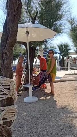 The video opens with a close-up of a tree trunk covered in rope. The camera then pans right, revealing a beach scene. In the foreground, a woman in a red and white striped swimsuit stands near a white umbrella and a table. To her right, a person in a green top and yellow shorts is folding a patterned fabric. Further back, a person in an orange shirt is bending down near the umbrella. A man in a white t-shirt and black shorts walks away from the camera towards a stone wall and some beach huts. The scene is characterized by gravel ground, trees, and beach umbrellas. The camera continues to pan right, showing more closed umbrellas and tables, and then moves to show a stone wall with steps leading up to beach huts. The video ends with a shot of a white railing on the right side of the frame and more beach huts in the background.