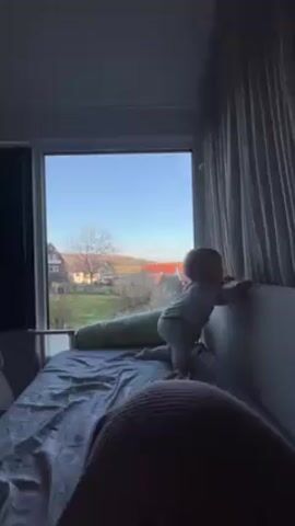 A baby in a patterned white onesie stands on a bed, looking out a window. The window offers a view of houses, trees, and a clear blue sky. The bed is covered with a gray sheet, and there is a long, green pillow behind the baby. A person is lying in the bed facing away from the baby. A curtain hangs to the right of the window, and a blind is visible on the left.