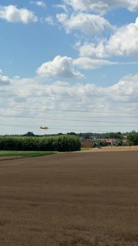 An orange airplane flies across a blue sky filled with white clouds. The plane is seen from the side and its tail is slightly angled upwards. Below the plane, there are power lines running horizontally across the frame. Further below the power lines is a landscape with a brown field in the foreground, followed by a line of green trees, and then a village with houses and a church steeple in the distance. The sun is out, casting light on the scene. The airplane appears to be flying at a relatively low altitude.