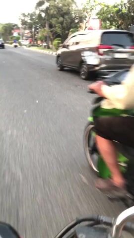 A man wearing a light-colored shirt and dark shorts rides a motorcycle on a paved road. He is wearing a green helmet. The motorcycle is black and green. The road is lined with trees and buildings. There are other vehicles and people visible in the background. The video appears to be filmed from another moving vehicle.