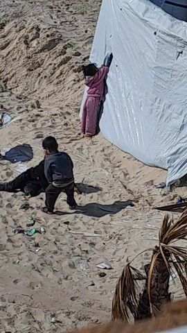 The aerial shot displays two individuals on a sandy surface, possibly a beach. One person, wearing dark clothing, is kneeling, apparently attending to something on the ground. The other person is lying under a white tarp, dressed in pink clothing.  A palm frond is also visible on the ground. The overall environment appears to be a makeshift camp or settlement.