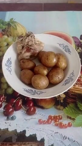 A plate of food containing chicken and potatoes sits on a table. The table covering has a fruit pattern. The food appears to be cooked. The plate is white with blue floral designs around the edge. The chicken is on the left side of the plate, and the potatoes are on the right. The potatoes are small and brown.